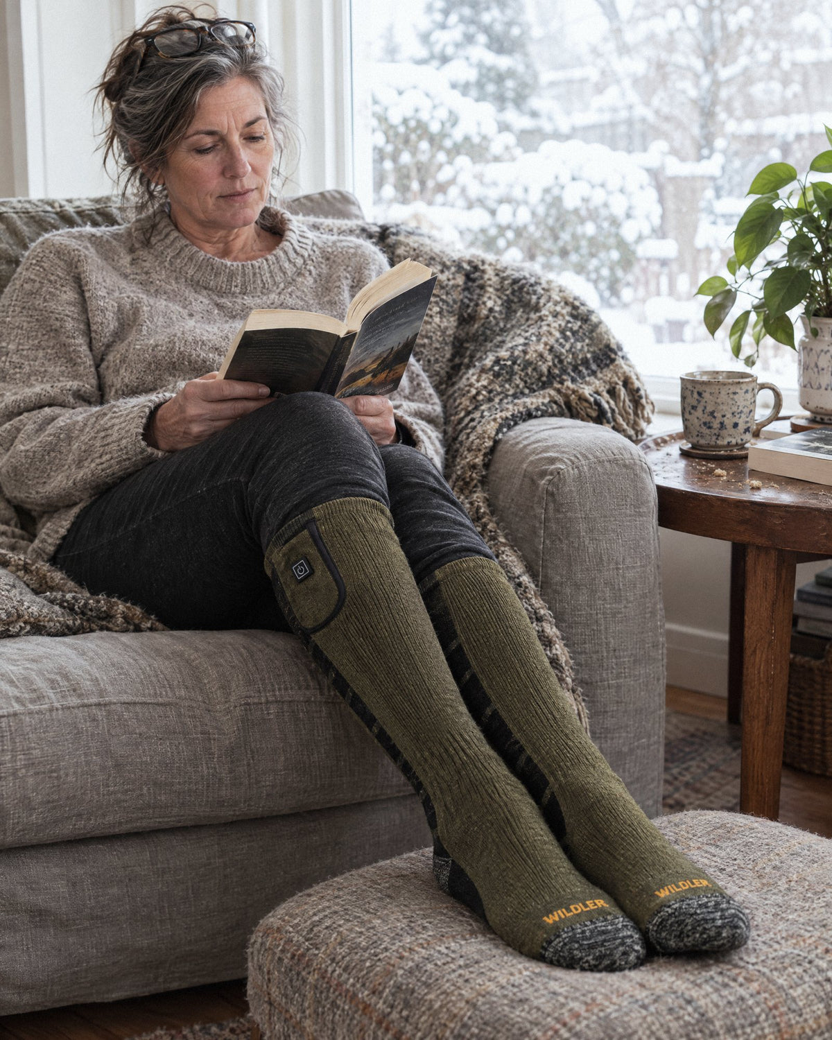Woman reading on a sofa with her feet up, wearing green Wildler Heated Socks